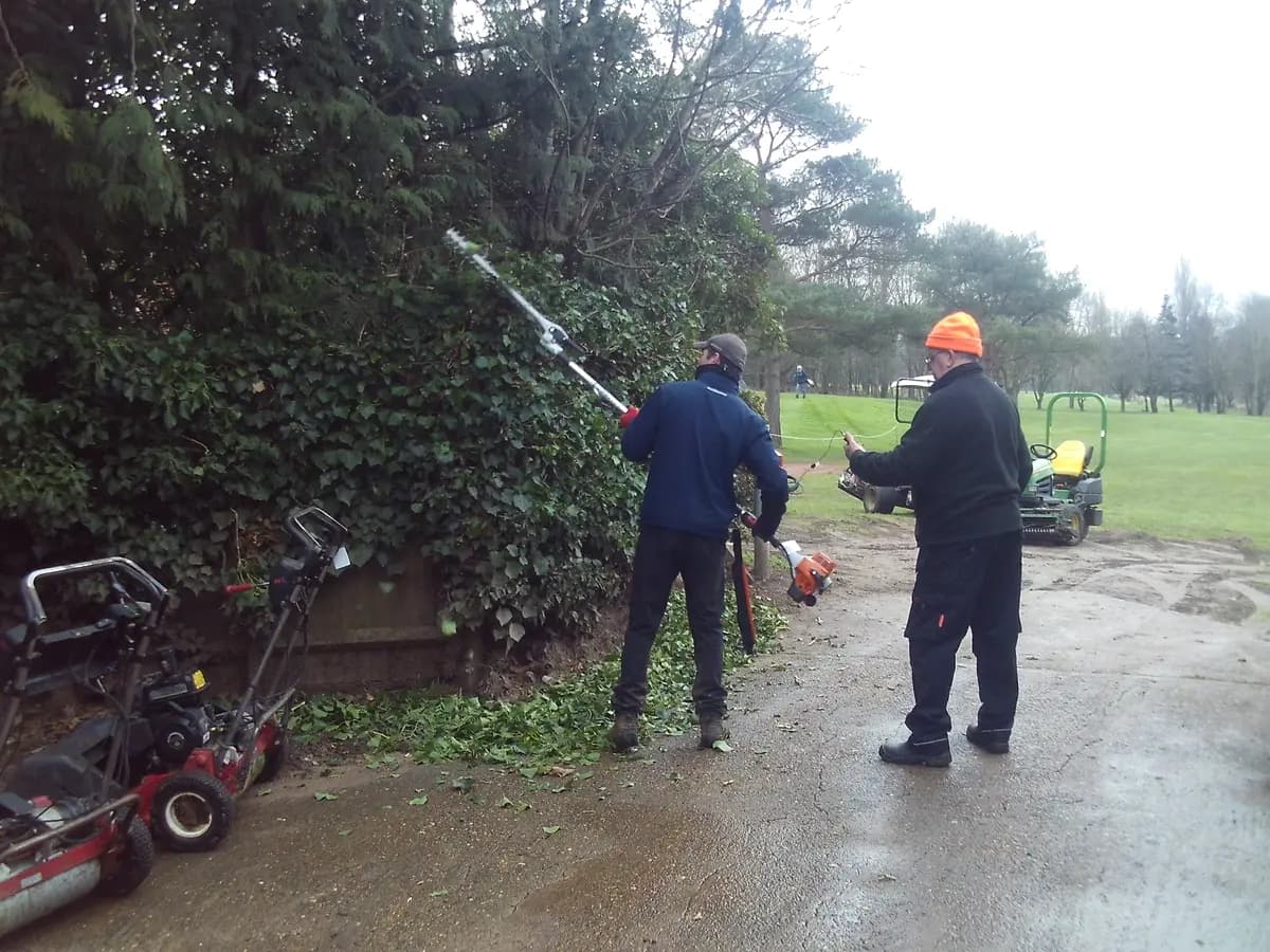Surveyor observing an operator using a pole hedge trimmer beside parked mowers at a golf club