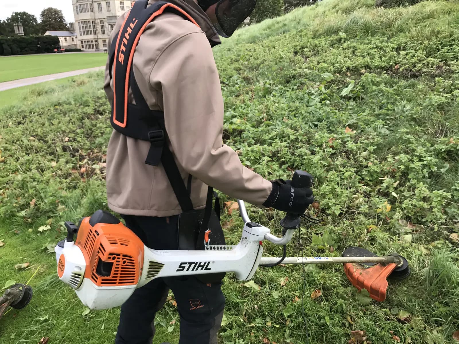 Operator in harness and face guard clearing a bank with a petrol brushcutter on estate grounds