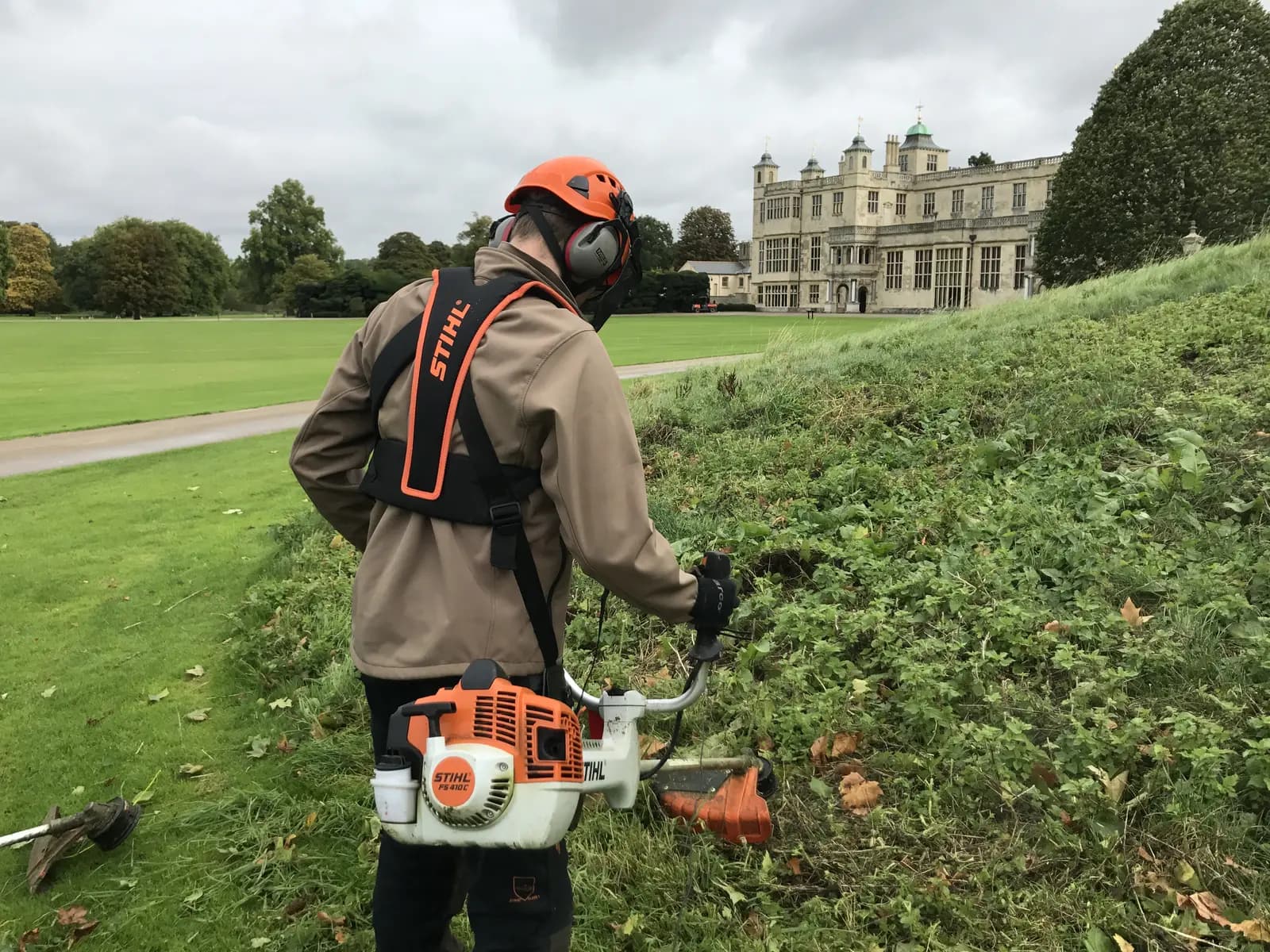 Operator clearing a grass bank with a brushcutter in the grounds of a historic estate