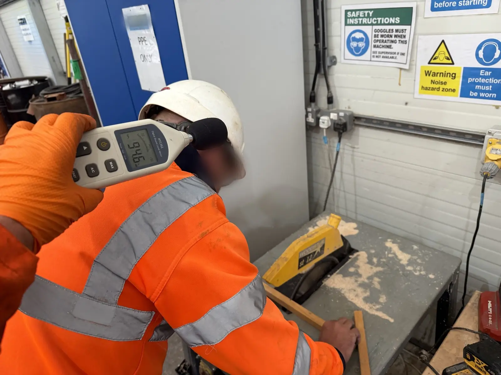 Sound level meter held at the operator's ear reading 94.6 dB while he works at a saw bench, beneath noise hazard and ear protection signage