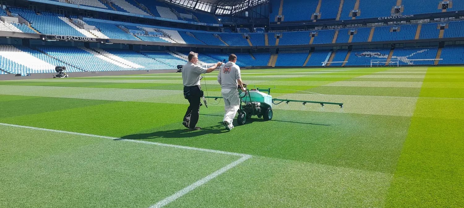 Noise assessment on a stadium pitch: a sound level meter held at the operator's ear while a pedestrian sprayer runs