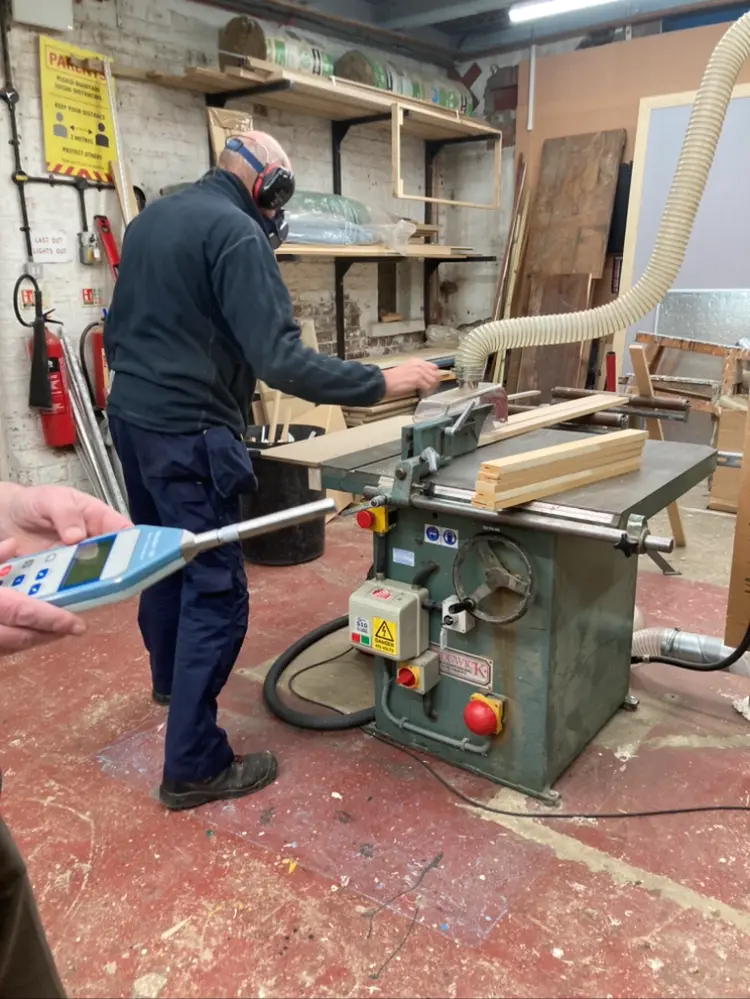 Sound level meter held towards a joiner in ear defenders working at a circular saw bench