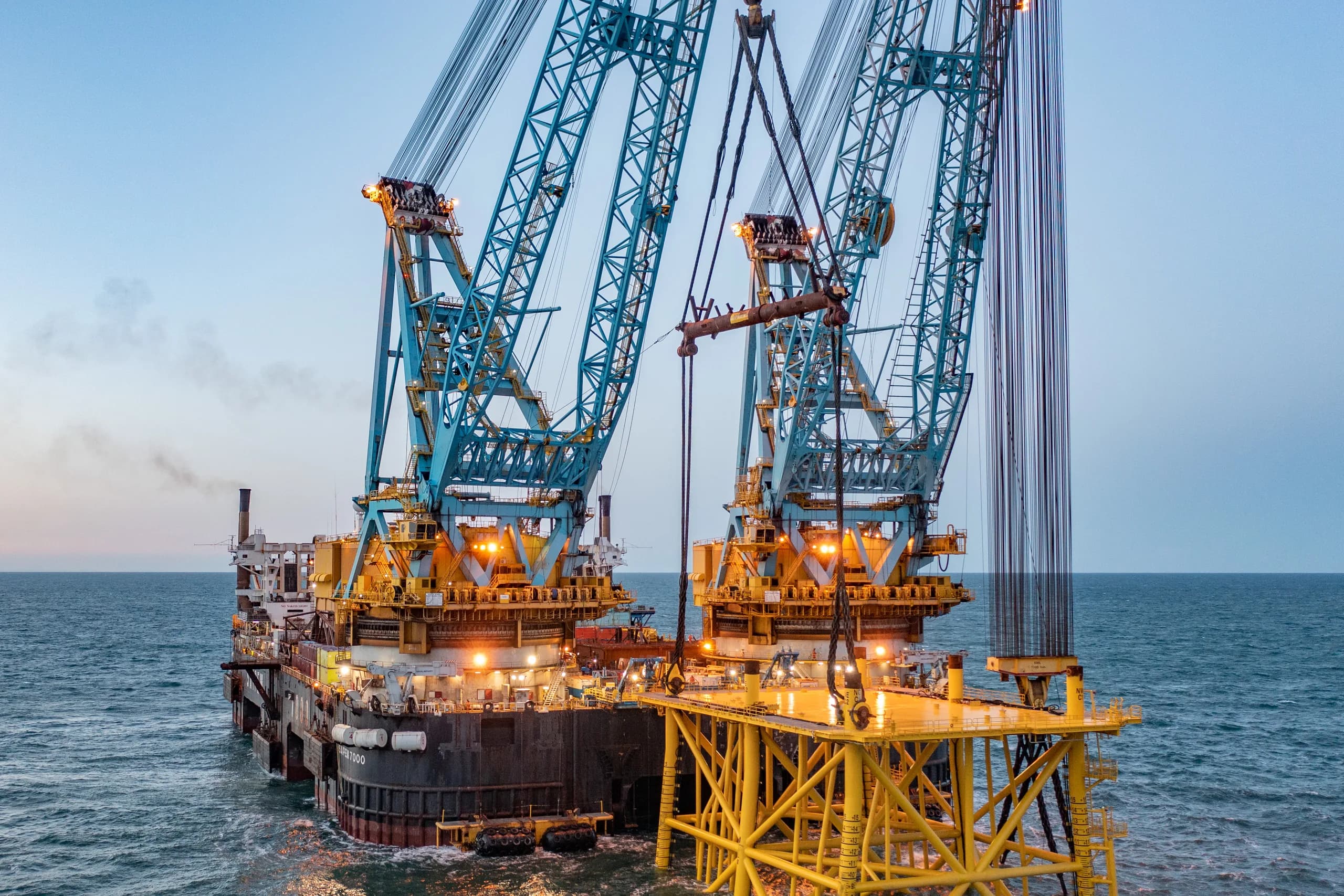Twin cranes of a heavy-lift vessel over a steel jacket offshore at dusk
