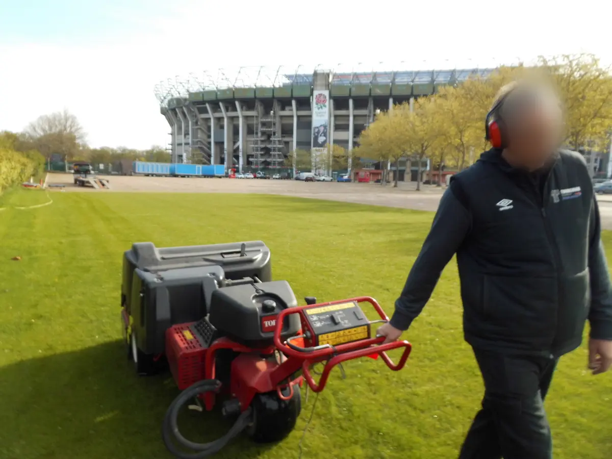 Operator in ear defenders wheeling a pedestrian aerator with the stadium behind