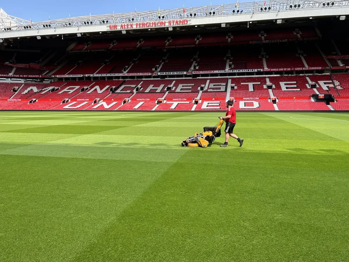 Groundsman walking a cylinder mower across a Premier League pitch beneath the stand