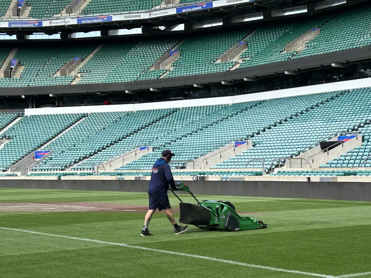 Operator pushing a pedestrian mower across a stadium pitch in front of the stands