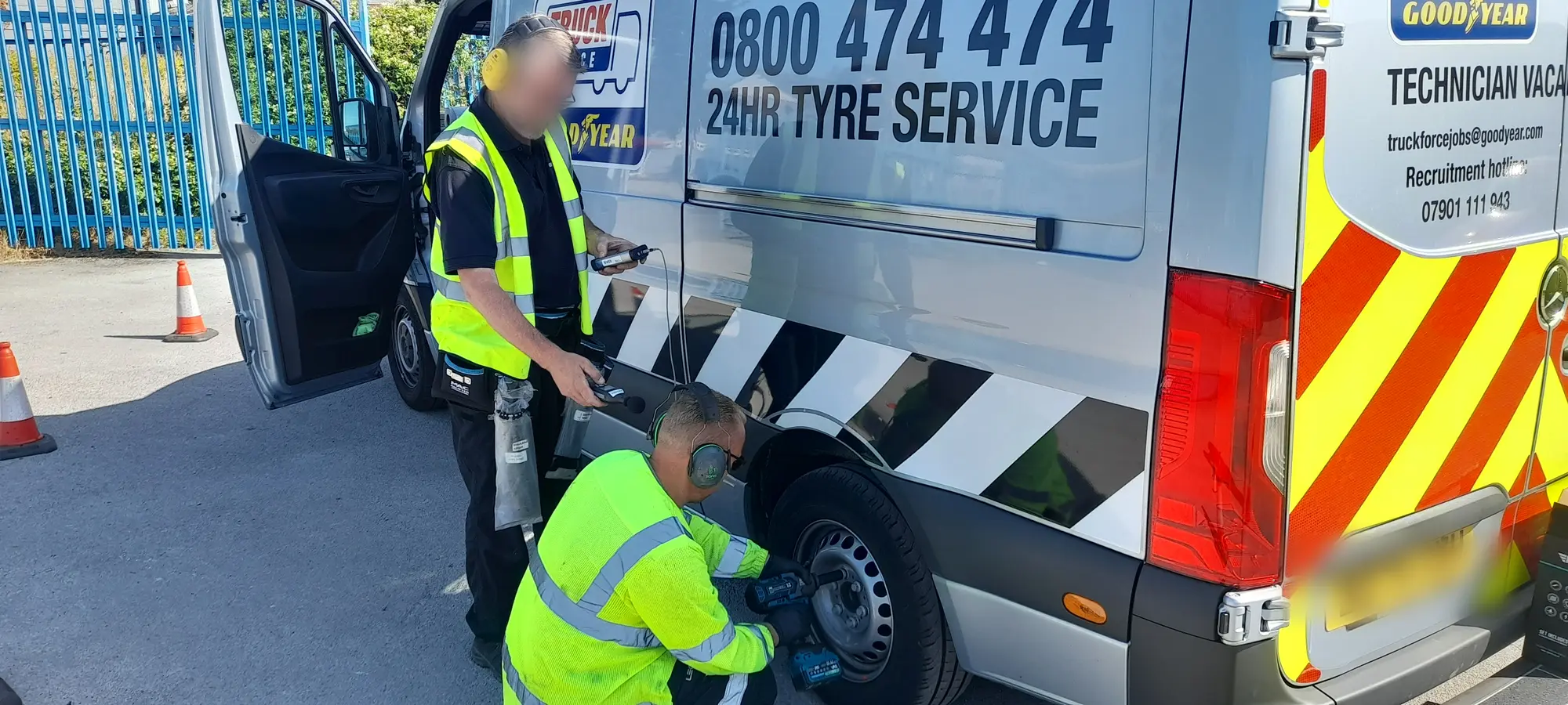 Surveyor holding a sound level meter over a technician in ear defenders running an impact wrench on a tyre-service van's wheel